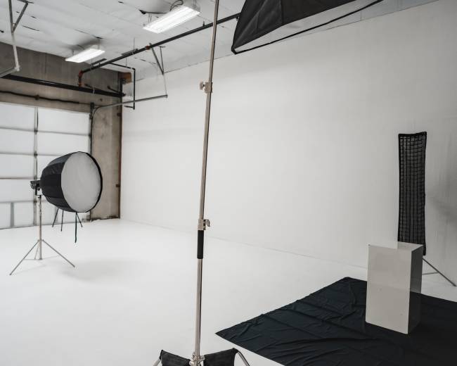 The image shows a photography studio with white walls, a large softbox light on a stand, a black backdrop on the floor, and an empty pedestal.