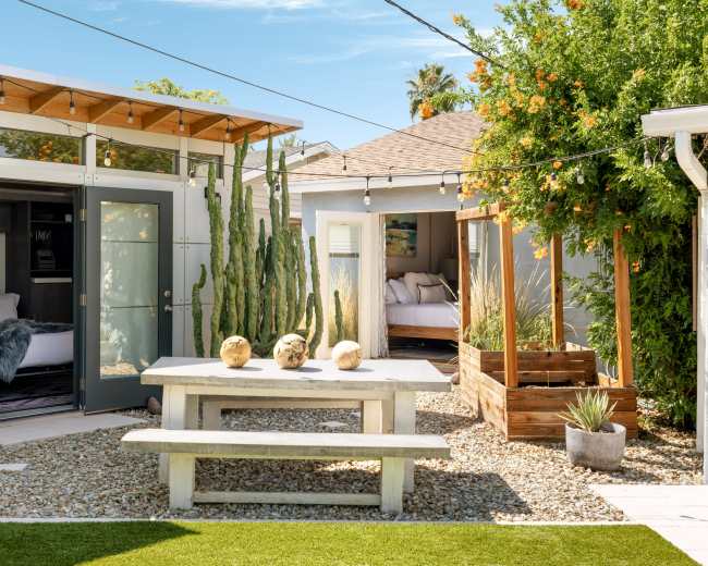 A modern outdoor courtyard features a wooden table with three decorative items, surrounded by cacti, gravel, and adjacent structures with large windows.