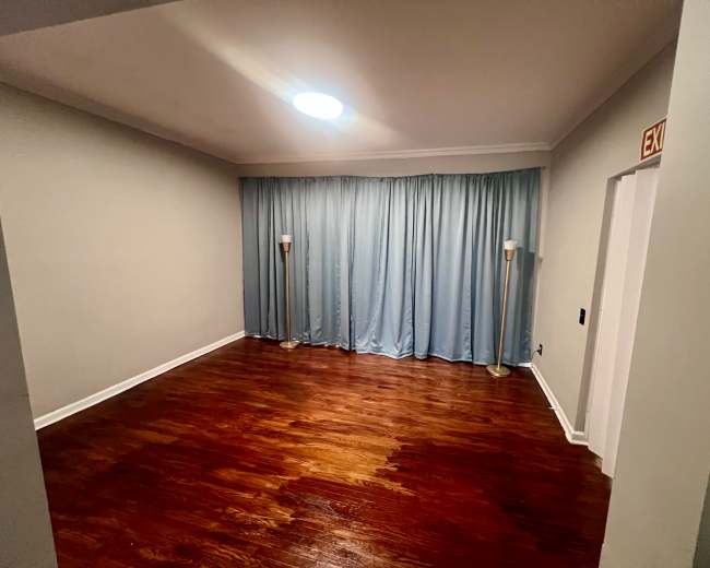 The image shows a sparsely furnished room with hardwood floors, light blue curtains, and two floor lamps near the curtains.