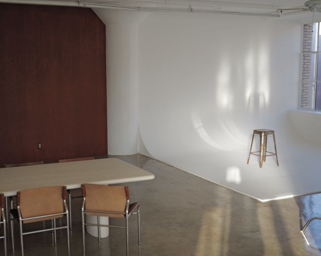 The room features a wooden wall on one side, a white curved backdrop, and a table with four chairs arranged around it.