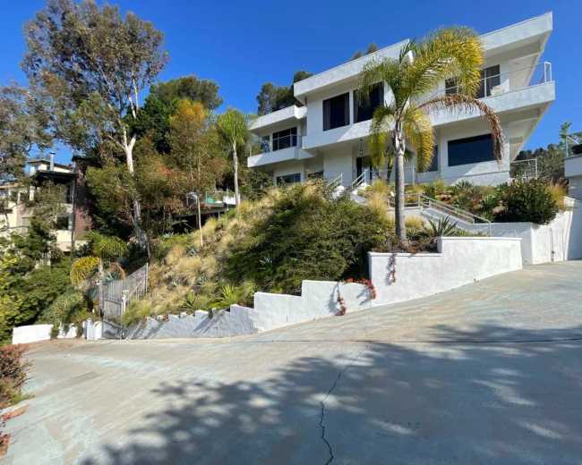 A modern house is situated on a hillside surrounded by greenery, with a winding driveway leading up to it.