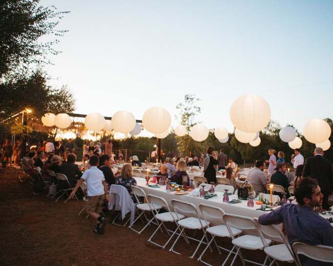 A large outdoor gathering features guests seated at long tables adorned with centerpieces, while white lanterns hang above in the evening sky.