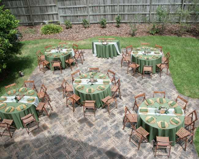 The image shows an outdoor event setup with several round tables covered in green linens, each surrounded by wooden chairs, arranged on a stone patio amidst a landscaped yard.