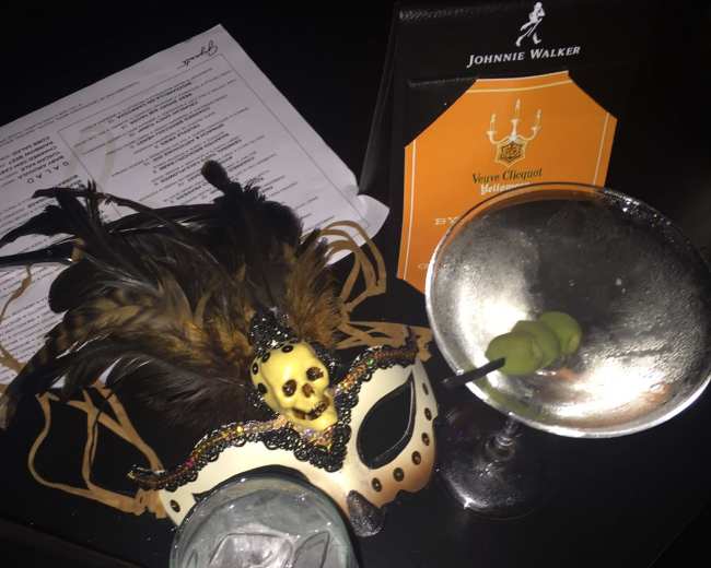 The image shows a masked carnival accessory alongside two cocktails on a table in a dimly lit setting.