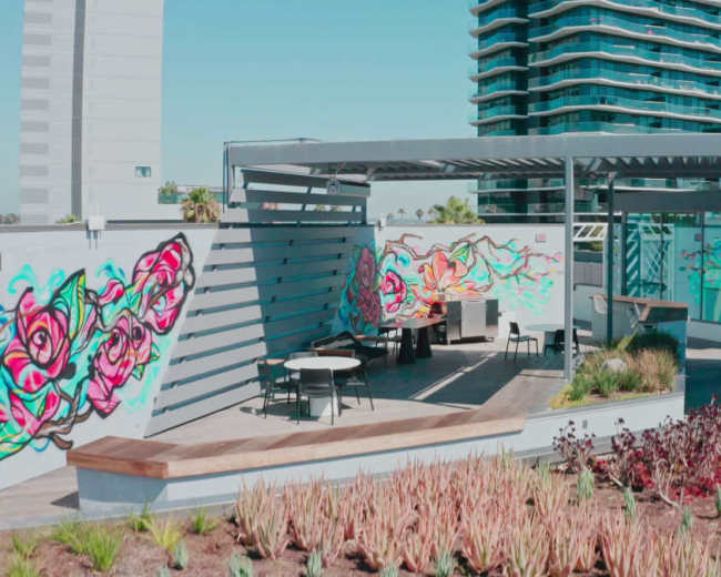 The image shows a rooftop space featuring vibrant mural art, modern seating, and landscaped greenery, with a view of a multistory building in the background.
