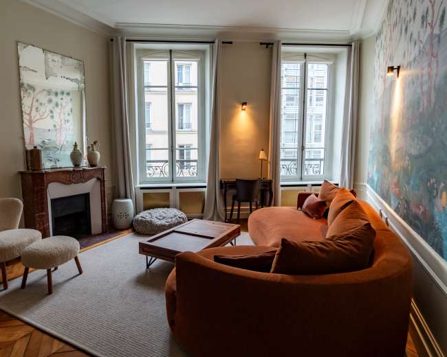 The image shows a cozy living room with large windows, featuring a curved orange sofa, a small table, a chair, and a decorative mirror on the wall.