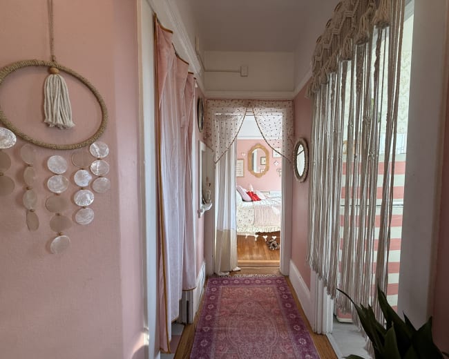 A pink hallway features draped curtains, decorative wall hangings, and a patterned rug leading to a visible room in the background.