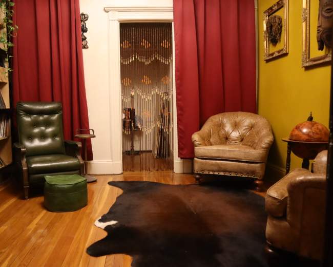 The image shows a cozy room with a record shelf on one side, two armchairs, and a cowhide rug on the wooden floor.