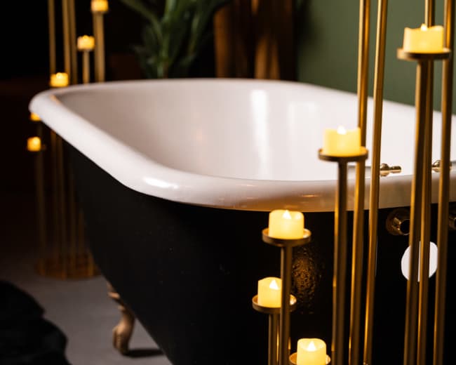 A black and white clawfoot bathtub is surrounded by slender gold candleholders with glowing candles, set against a green wall.