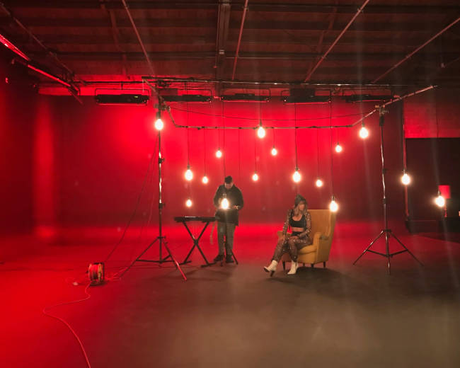A woman sits in a yellow chair under hanging lights while a man stands at a keyboard in a red-lit studio.