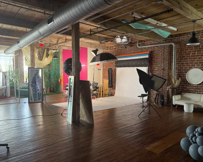 The image shows a spacious photography studio with various backdrops, lighting equipment, and creative props arranged around a hardwood floor.