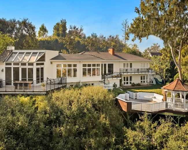 A large, modern white house with multiple deck areas is situated amidst lush greenery and trees.