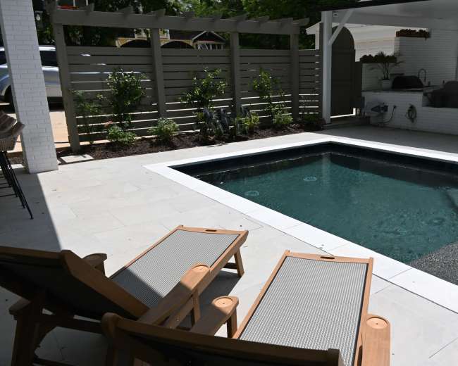 The image shows a modern outdoor space featuring a swimming pool surrounded by patio tiles and two wooden lounge chairs.