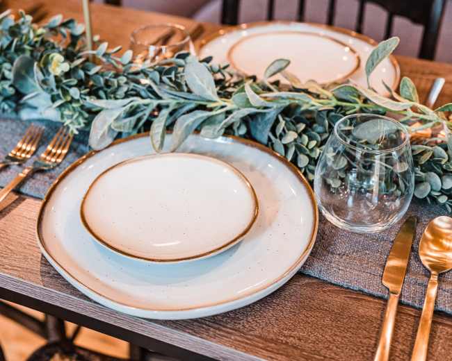 A dining table is set with a beige table runner, two plates, cutlery, and glassware, adorned with greenery along the center.