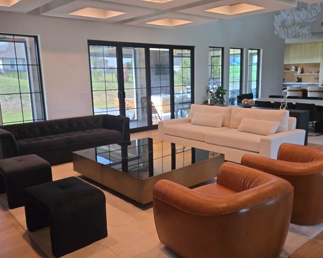 The image shows a modern living room with two black sofas, a white sofa, an elegant glass coffee table, and brown leather armchairs, all arranged around a central seating area.