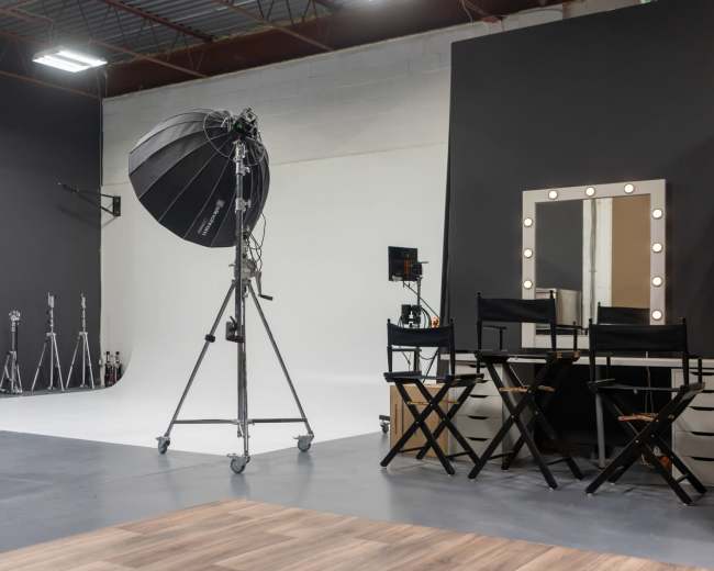 The image shows a photography studio equipped with lighting equipment, black walls, a white backdrop, and director's chairs arranged in front of a mirror with lights.