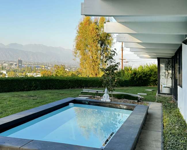 The image shows a modern backyard with a swimming pool, surrounded by grass and overlooking a mountainous landscape.