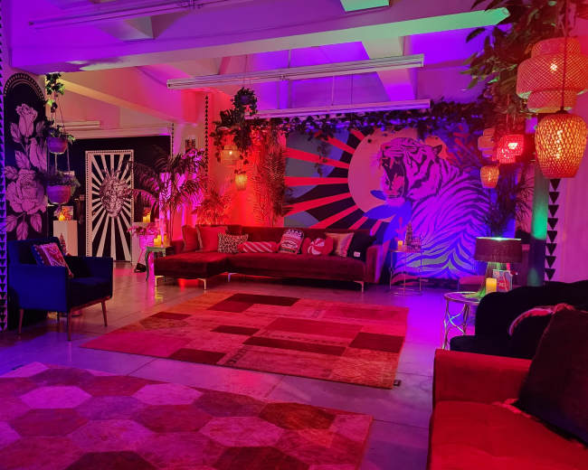 The image features a vibrant and colorful lounge area with bold wall art, mixed seating arrangements, and decorative lighting in a modern interior space.