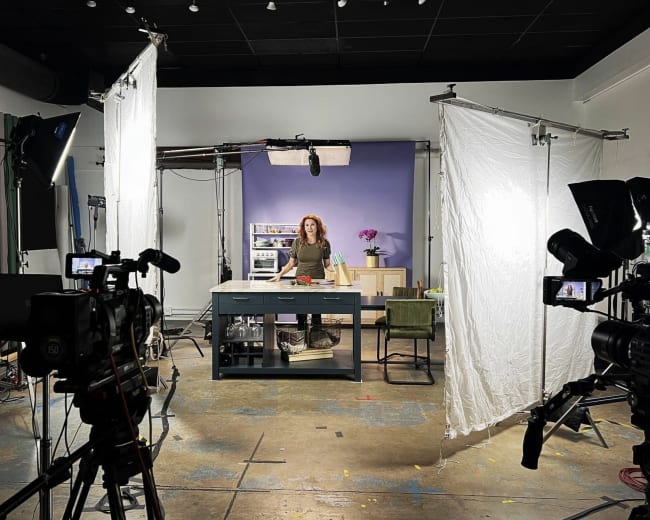 A video production set features a woman standing behind a table surrounded by cameras, lights, and backdrop elements.