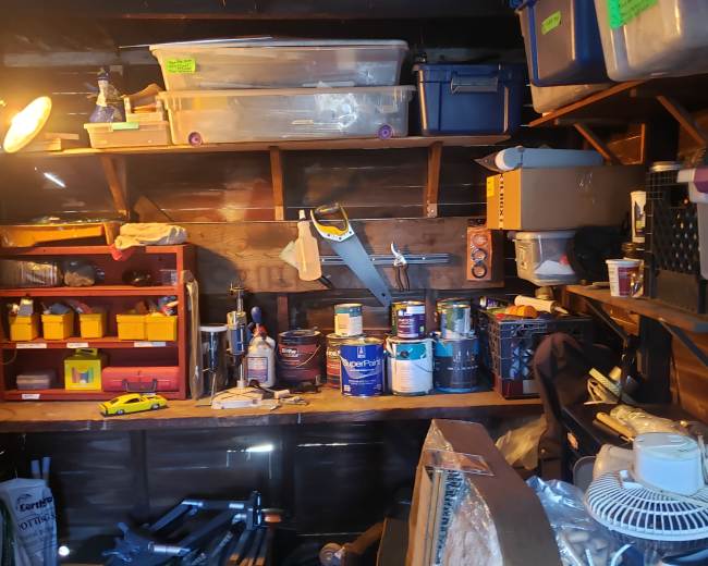 A cluttered storage shed filled with containers, tools, and paint cans organized on shelves and tables.