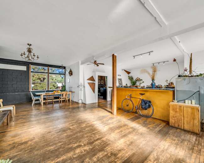 The image shows a spacious interior with hardwood flooring, a dining area with chairs and a table, and a kitchen counter featuring a bicycle parked beside it.