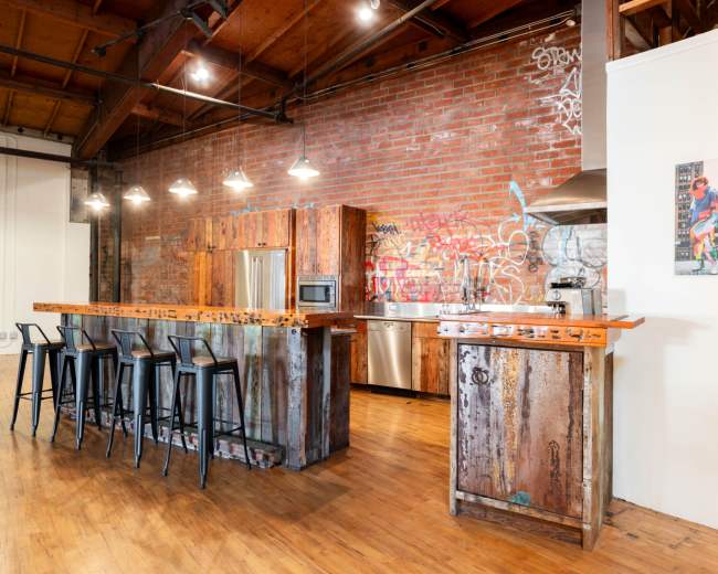 The image shows a spacious kitchen with a rustic wood and metal island, industrial-style stools, stainless steel appliances, and exposed brick walls featuring graffiti.