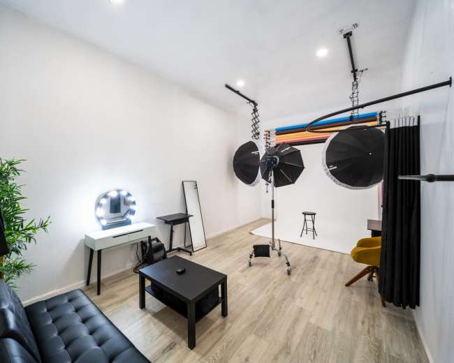 The image shows a photography studio with lighting equipment, a stool, a mirror, and a sofa arranged in a well-lit, minimalist space.