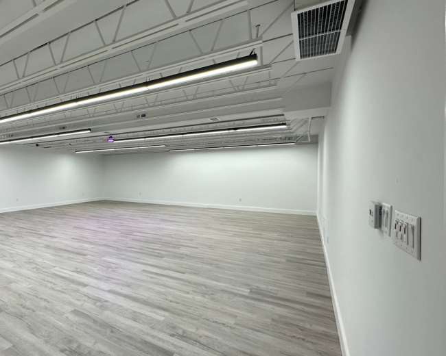 The image shows an empty room with white walls, a light wooden floor, and overhead lighting fixtures.