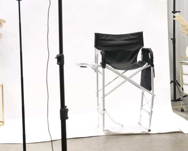 A black director's chair is positioned in front of a plain white backdrop, illuminated by a mounted light source on a stand.