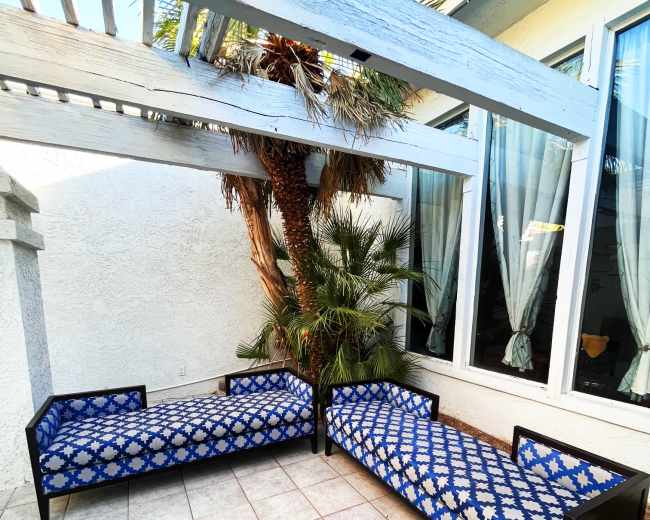 Two lounge chairs with blue patterned cushions are positioned under a wooden pergola beside large windows and tropical plants.