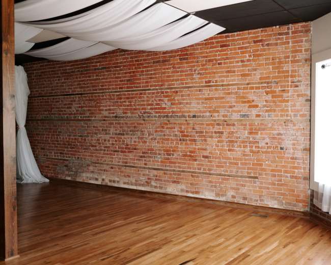 The image shows an interior space featuring a brick wall, hardwood flooring, and a large window with white curtains.