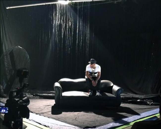 A person sits alone on a black couch in a dimly lit studio with a backdrop of hanging strands and a camera setup nearby.