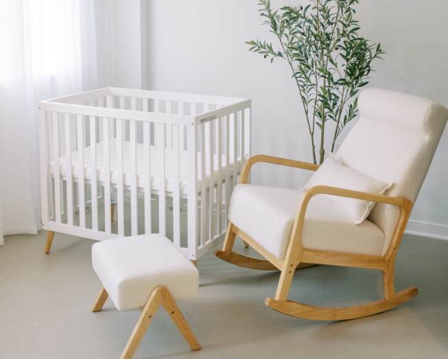 The image shows a bright nursery featuring a white crib, a rocking chair, a footrest, and a potted plant near a large window.