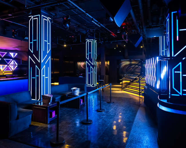 The image shows a modern club interior featuring illuminated columns, a bar area with geometric designs, and a sleek, polished floor.