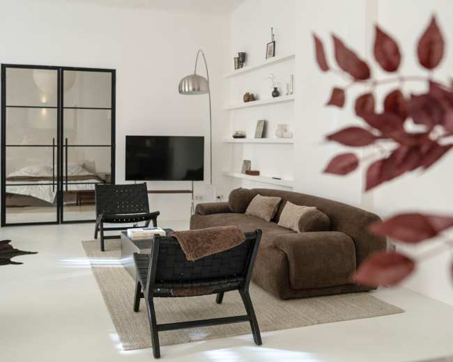 The image shows a modern, minimalist living room with a brown sofa, two black chairs, a television, and a shelving unit against a white wall.