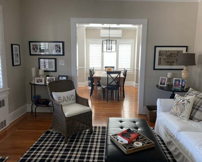A living room features a white sofa, a black ottoman on a checkered rug, and a view into a dining area with a wooden table and window shutters.