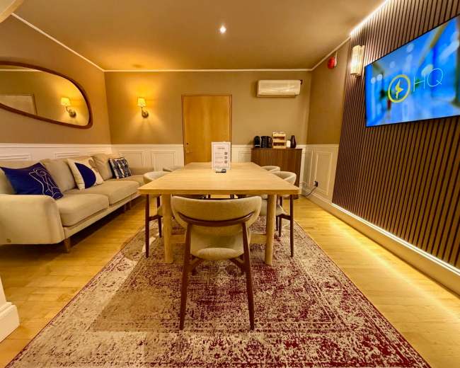 A modern meeting room features a long wooden table surrounded by chairs, a sofa, and an illuminated wall with a screen displaying a logo.