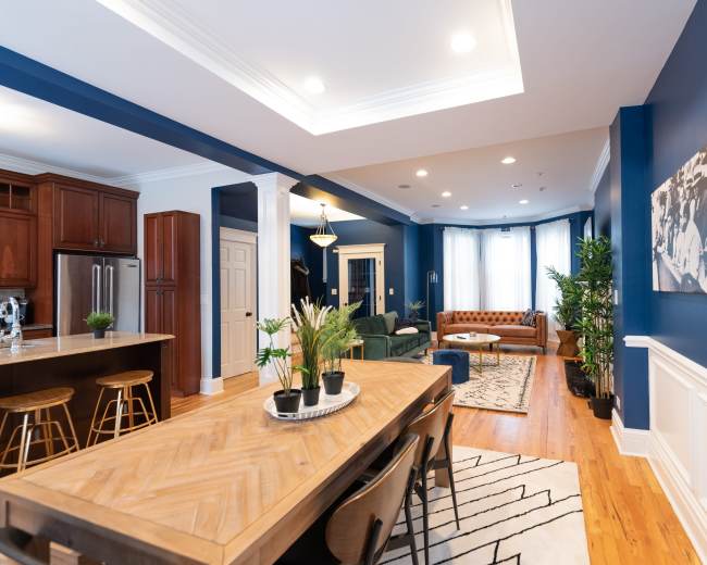 A spacious living and dining area features a wooden dining table, modern bar stools, a green sofa, and brown leather seating, with blue walls and large windows allowing natural light.