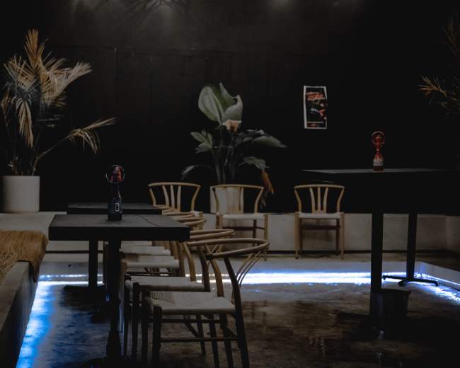 A dimly lit interior of a modern cafe features wooden tables and chairs, with potted plants in the background and a strip of blue LED lighting along the floor.