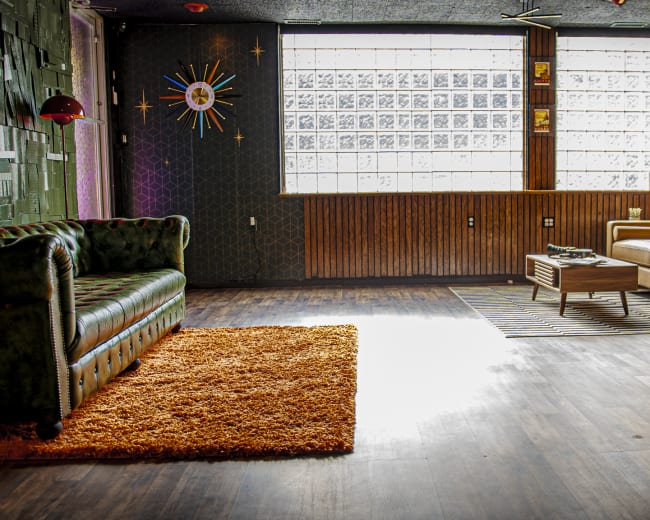 The image shows a spacious room with a green velvet sofa, a brown leather couch, and a wooden coffee table, featuring a large window with glass blocks that allows natural light to enter.