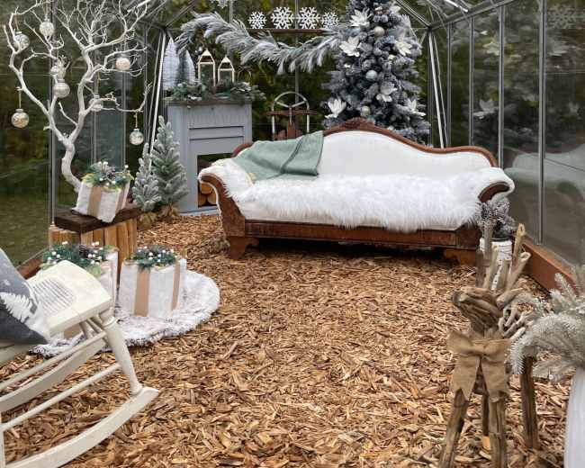 The image shows a cozy greenhouse decorated for winter with a white sofa, a Christmas tree, and snowflake ornaments.
