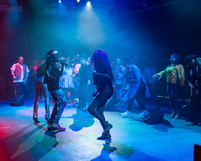 A performer sings on stage while audience members dance and engage with the music in a dimly lit venue.