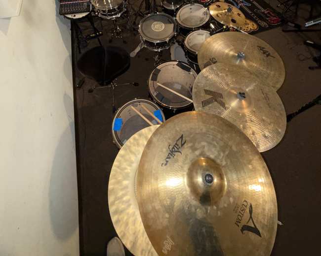 The image shows a rehearsal space with a large drum set, various percussion instruments, electronic equipment, and a microphone setup.