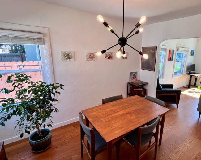 A dining area features a wooden table surrounded by chairs, with a chandelier overhead and a potted plant, leading to a living room visible through an archway.