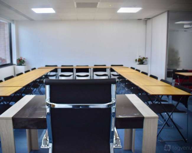 A large conference room with multiple rectangular tables arranged in a U-shape, surrounded by black folding chairs and bright, natural light coming through the windows.