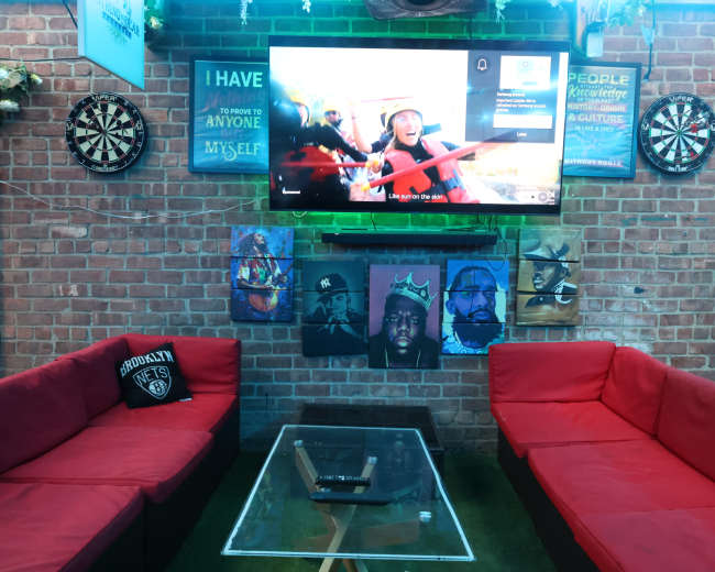 The image shows a cozy lounge area with red couches, a glass coffee table, and a wall adorned with artwork and a television displaying a program.
