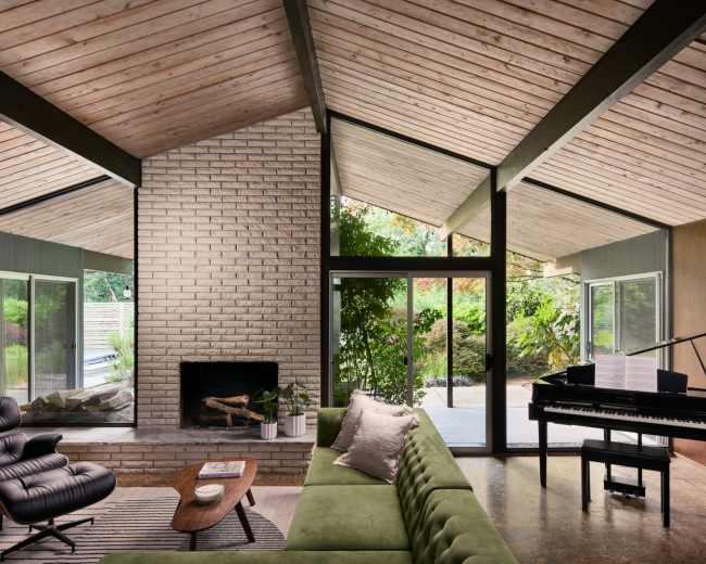 The image shows a modern living room with a green sectional sofa, a brick fireplace, large windows, and a piano placed near an indoor plant.