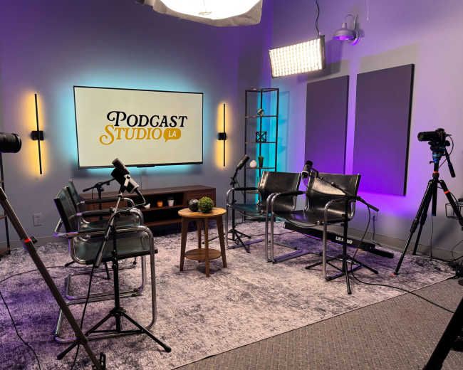 The image shows a podcast studio with two chairs, a small wooden table, and multiple cameras set up in front of a large screen displaying the text "PODCAST STUDIO LA."