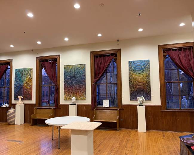 The image shows an art gallery with six large abstract paintings displayed on the walls, framed by windows dressed with red curtains, and two small tables in the center.