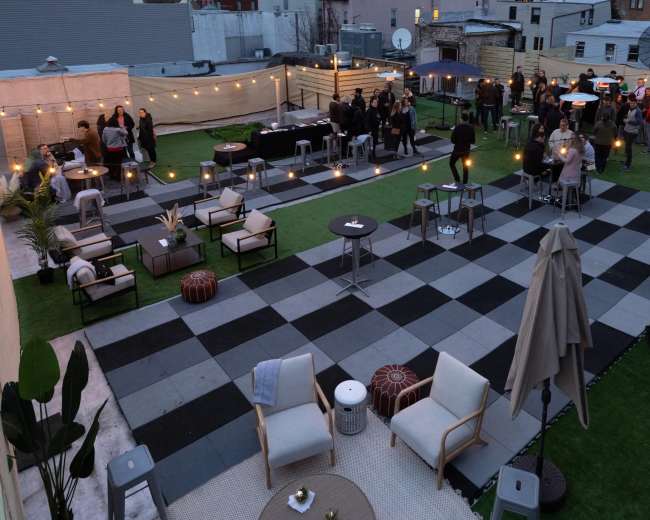 The image shows a rooftop event space with a checkerboard pattern of black and white tiles, seating areas, and string lights illuminating a gathering of people.